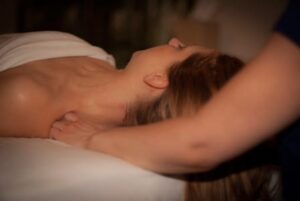 The left side profile of a woman's face with white skin and light brown, straight hair resting face up on a massage table with white sheets with a knuckles of a female massage therapist pressing into her left trapezius muscle.