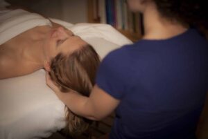 Woman with white skin and light brown, straight, medium lengthed hair, laying face up on a massage table with a white sheet covered her chest is receiving craniosacral therapy by a female therapist with white skin, wearing a deep blue t-shirt and resting her hands under the client's base of skull. The back of the therapist is seen and not the front.