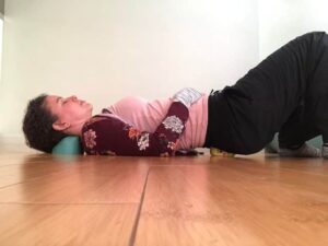 Woman wearing a pink tank top, black pants and maroon long sleeve top with flowers printed on it, laying on her back on top of a seafoam green craniocradle cnd yellow 4" myofascial release therapy ball with both knees bent and feet flat on a light brown wooden floor.