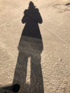 Large shadow of a woman wearing a winter hat and coat on a beige, concrete sidewalk on a very sunny day.