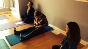 Three women seated on the floor. Two women sitting on one purple and one blue yoga mats, two women are crossed legged and one woman has two legs stretched out in front of her looking down as she zips up her sweatshirt jacket. The floor is light brown wood and the wall is painted grey with white floor board paneling.