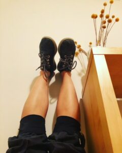 Two feet in grey tied sneakers with bare calves and black knee-length pants resting up on a wall next to a light brown bookshelf with a white vase filled with yellow circular flowers on top of the shelf.