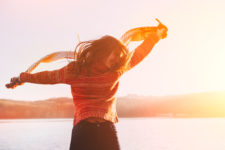 Woman wearing orange sweater with arms stretched out at her sides holding a scarf that's blowing in th wind with a lake in the background