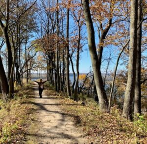 Long gravel path with trees lining the path with autumn colored leaves of orange, green and gold. Some dried leaves are on the ground next to the path and a female standing in the middle of the path wearing blue pants, white shirt and black cardigan with arms stretched out to her side up in the air. Blue skies and blue lake surrounded by a tree line in the background.