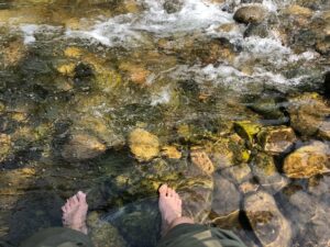 A pair of bare feet soaking in a riverbed in the foreground, on top of several medium sized brown rocks covered in green algae and white colored rushing water in the back of the photo.