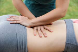 An upclose image of a cross hand myofascial release technique on female sacrum and lumber spine. Therapist wearing a green shirt and client wearing a light grey sports bra and yoga pants.