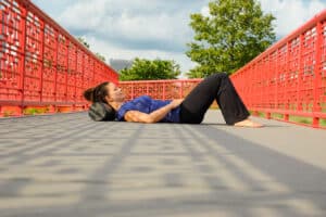 Female laying down on back and resting neck on black foam roller with red railing bridge and trees in the background