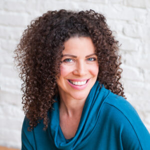 Heather Fraelick wearing a teal scoop neck sweater seated in front of a white brick wall with shoulder length curly hair down and big smile.