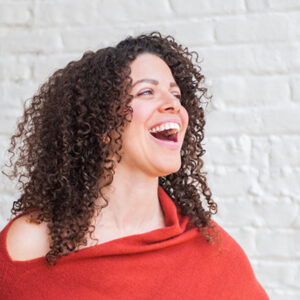 Side profile image of Heather Fraelick, LMT laughing and wearing orange blouse in front of a white brick wall.