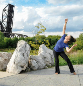 Female dancer wearing a bright blue top and black pants, standing with her torso stretched over her left leg, her head looking up over her right shoulder to the blue cloudy sky and right arm stretched up towards the clouds. She's dancing on cement ground with large rocks behind her to her right, green grass and trees int eh background with an industrial bridge drawn up to the back left of the photo.