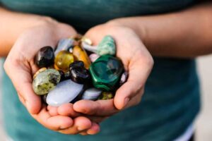 Two cupped hands in front of a person wearing a green top, holding two palm fulls of crystals of various colors from light blue to dark green, yellow, pink and black.