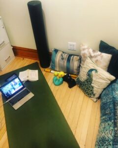 Green yoga mat laid out on an angle to the left on a light wood floor with a rose gold Apple laptop computer and open book face down resting on the mat. One 36" black foam roller resting vertically against the wall and various massage balls and sticks laying next to 5 teal and gold pillows laying on the floor next to it.