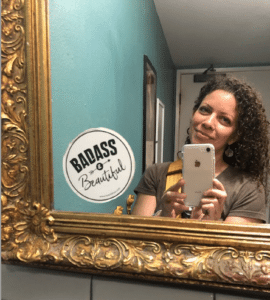 Carmel brown skin woman with dark brown curly hair looking into an antique gold framed mirror while holding her iPhone to take a selfie, with a teal painted wall in the back ground and a white circular sticker on the mirror that says, "Badass & Beautiful" in black letters.