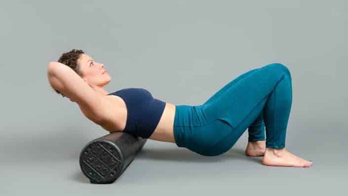 Heather Fraelick barefoot wearing a navy blue top and teal pants laying upper back horizontally on a black foam roller with her hair pulled back, hands clasped behind the base of her skull and slight smile on her face.