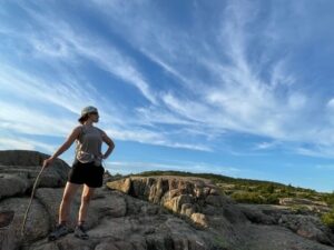 Woman holding a walking stick, looking away from the camera to her left and wearing a baseball cap, black shorts, tan tank top, hiking boots and backpack striped to her back and waist, standing on top of a mountain with a big blue sky in the background and grassy field to the right of her and large light brown rocks underneath her.