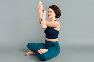 Woman wearing a dark blue sports bra and teal tights sitting cross legged seated eagle arm yoga posture, looking at the camera with a closed mouth smile and twinkle in her eye. Simple grey background.