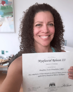 Heather Fraelick holding her MFR III certification of completion with a huge smile on her face and long brown curly hair draping over her shoulders with a vibrant multi-colored painting in the background