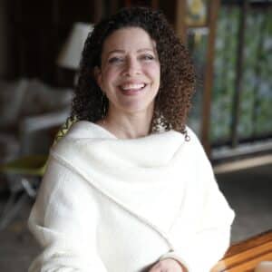 Woman with brown curly hair, wearing a white sweater sitting at a wooden desk smiling at the camera. Her arms are resting relaxed on the desk and there's a green stained glass window behind her, a lamp with a white lampshade and a couch behind her as well.