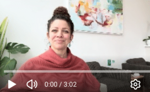 Brunette woman with her curly hair in a high bun, smiling with mouth closed, wearing an orange sweater seated in a room with white walls, a brightly rainbow colored painting in the background and two dark grey cushiony chairs.