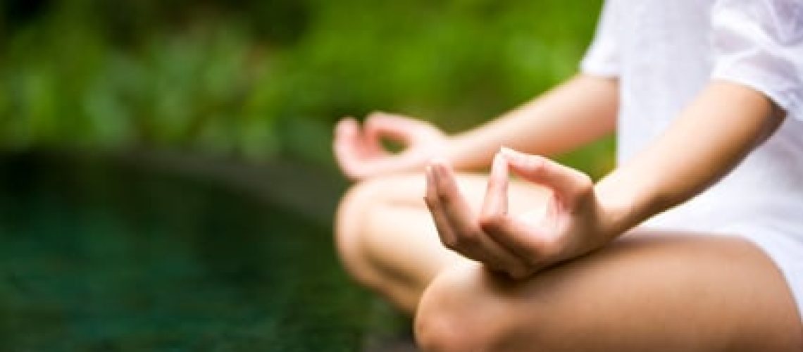 White woman seated by a lake wearing a white shirt and shorts with both legs crossed and hands resting on her thighs with thumb and forefinger touching in a meditative position.
