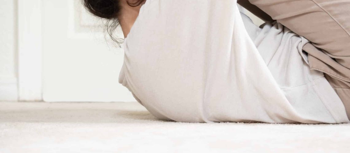 The side profile of a brown haired woman wearing a white shirt and khaki pants hugging her knees into her chest while laying on a white floor with white wall in the background.